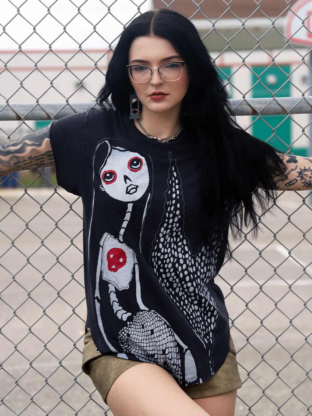 Model wearing a black t-shirt with a Mermaid graphic design, leaning against a chain-link fence.