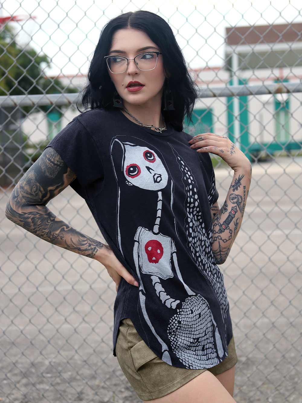 Woman wearing a black t-shirt with a skeleton mermaid design in front of a chain-link fence.