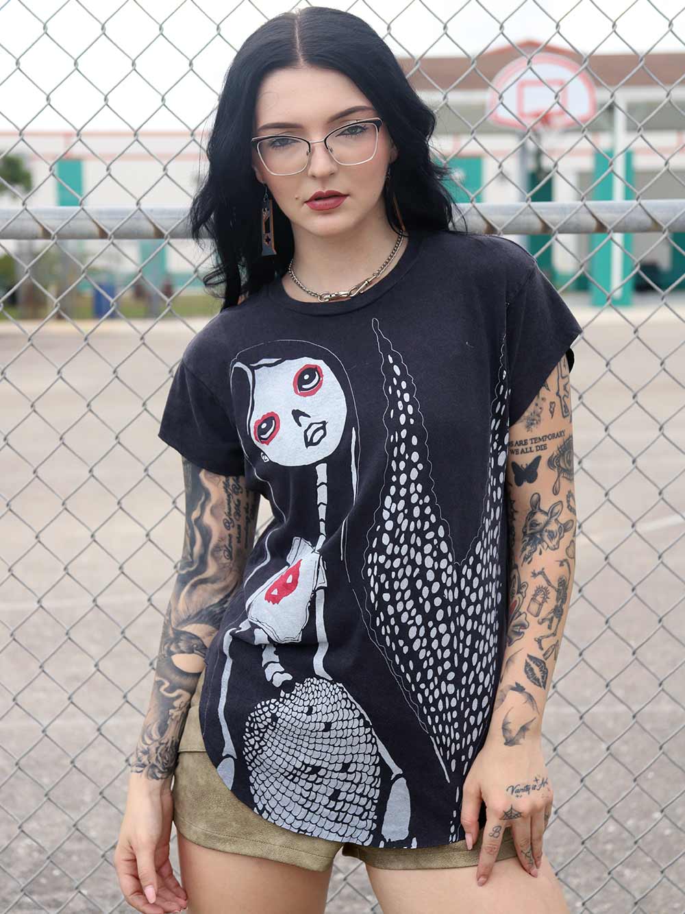 Person wearing a black t-shirt with a Mermaid graphic design, standing in front of a chain-link fence.