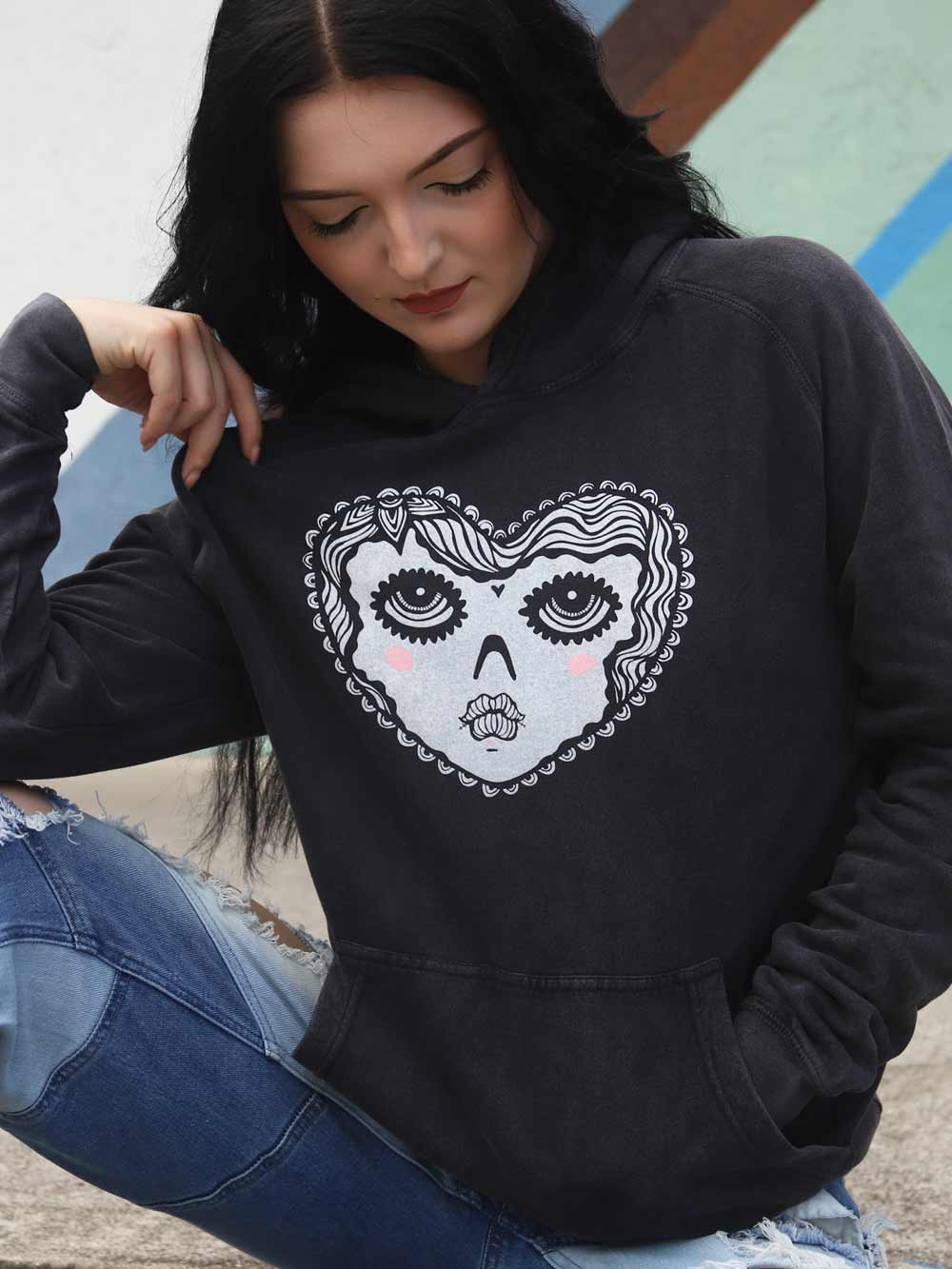 Woman wearing a black hoodie with a graphic heart face design, sitting on a concrete surface.
