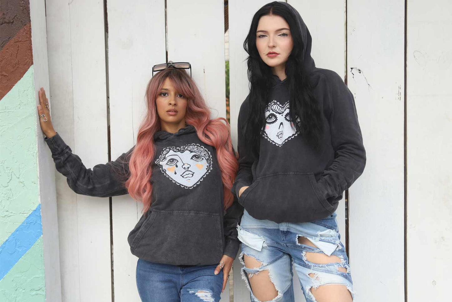 Two women wearing black hoodies with graphic designs against a white wall.