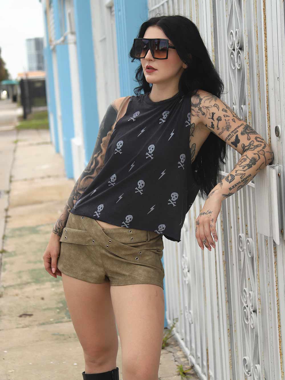 Woman with tattoos wearing a black sleeveless top with skull & lightning bolt patterns, leaning against a white metal gate.