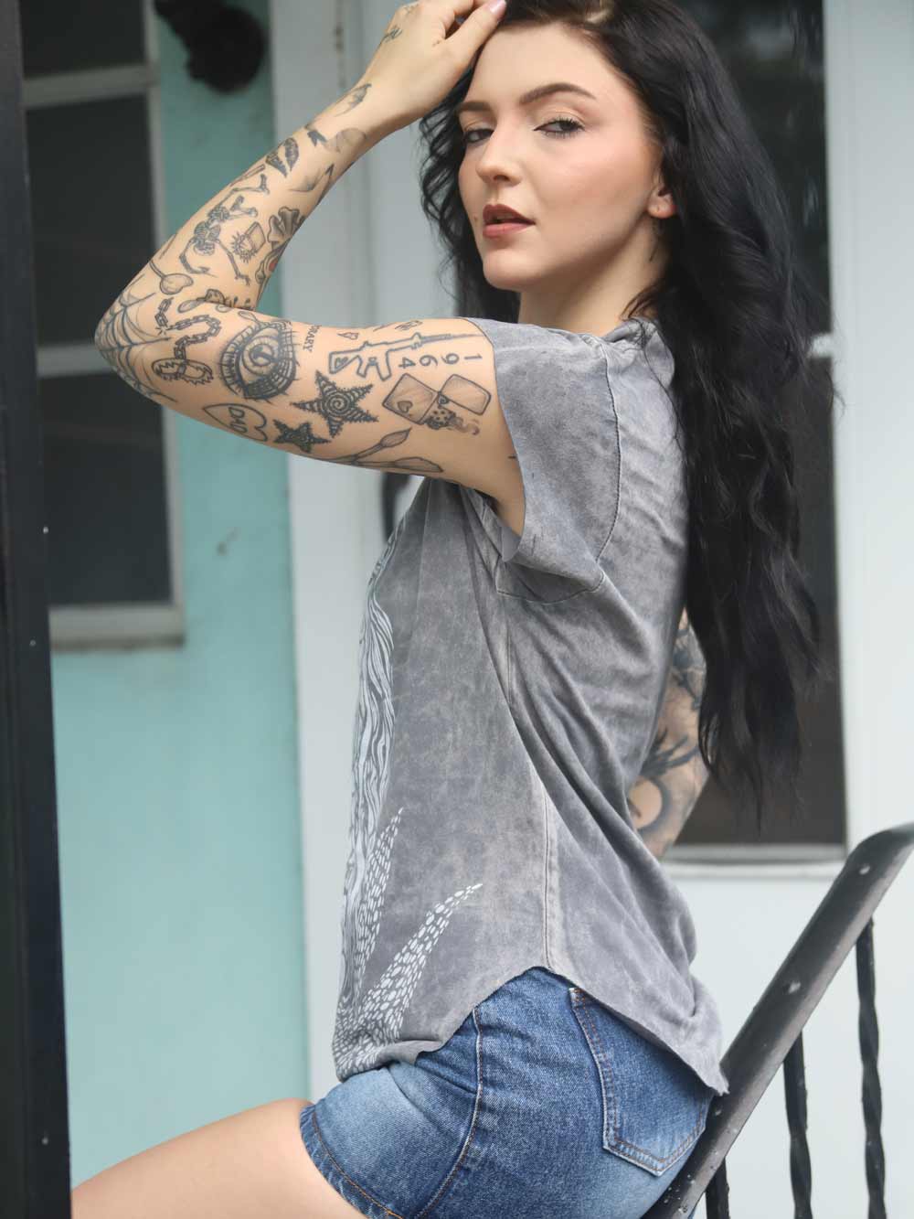 Woman with tattoos wearing a gray mermaid t-shirt and denim shorts outdoors.