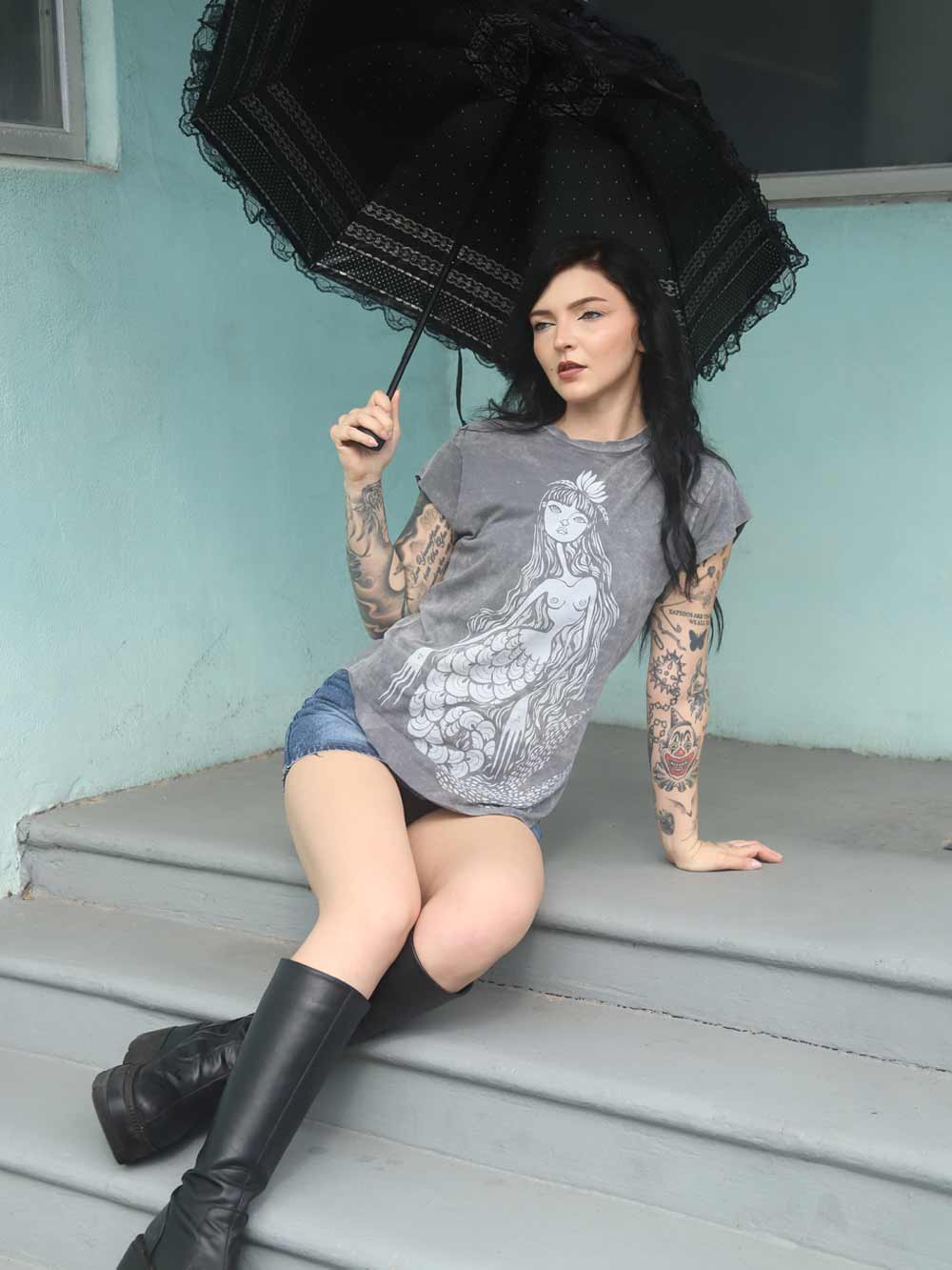 Woman sitting on steps with a mermaid design shirt, holding a black umbrella against a light blue wall.