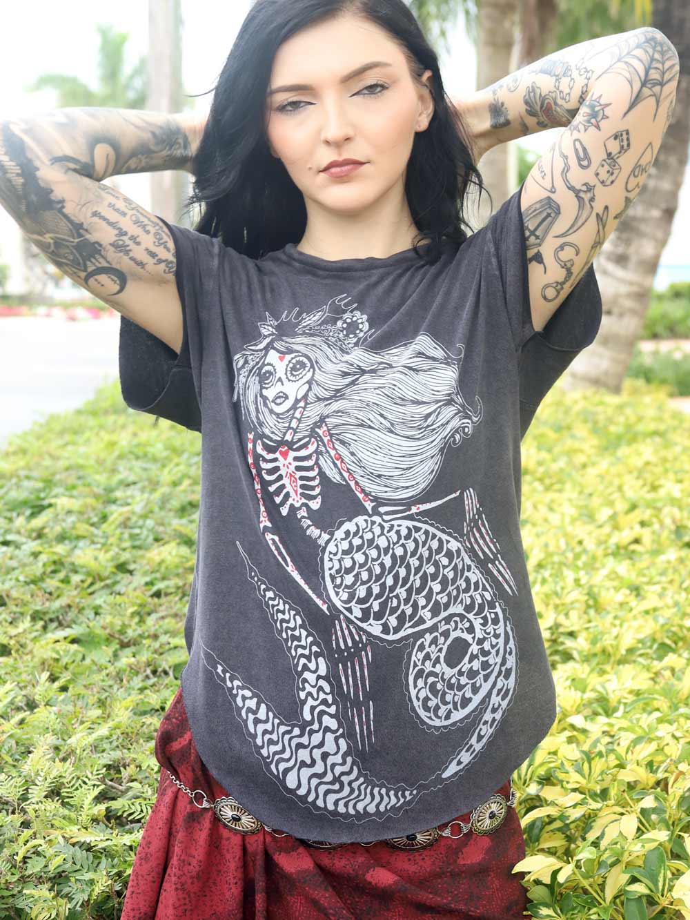 Model wearing a dark gray t-shirt with a skeleton mermaid design, standing outdoors with greenery in the background.