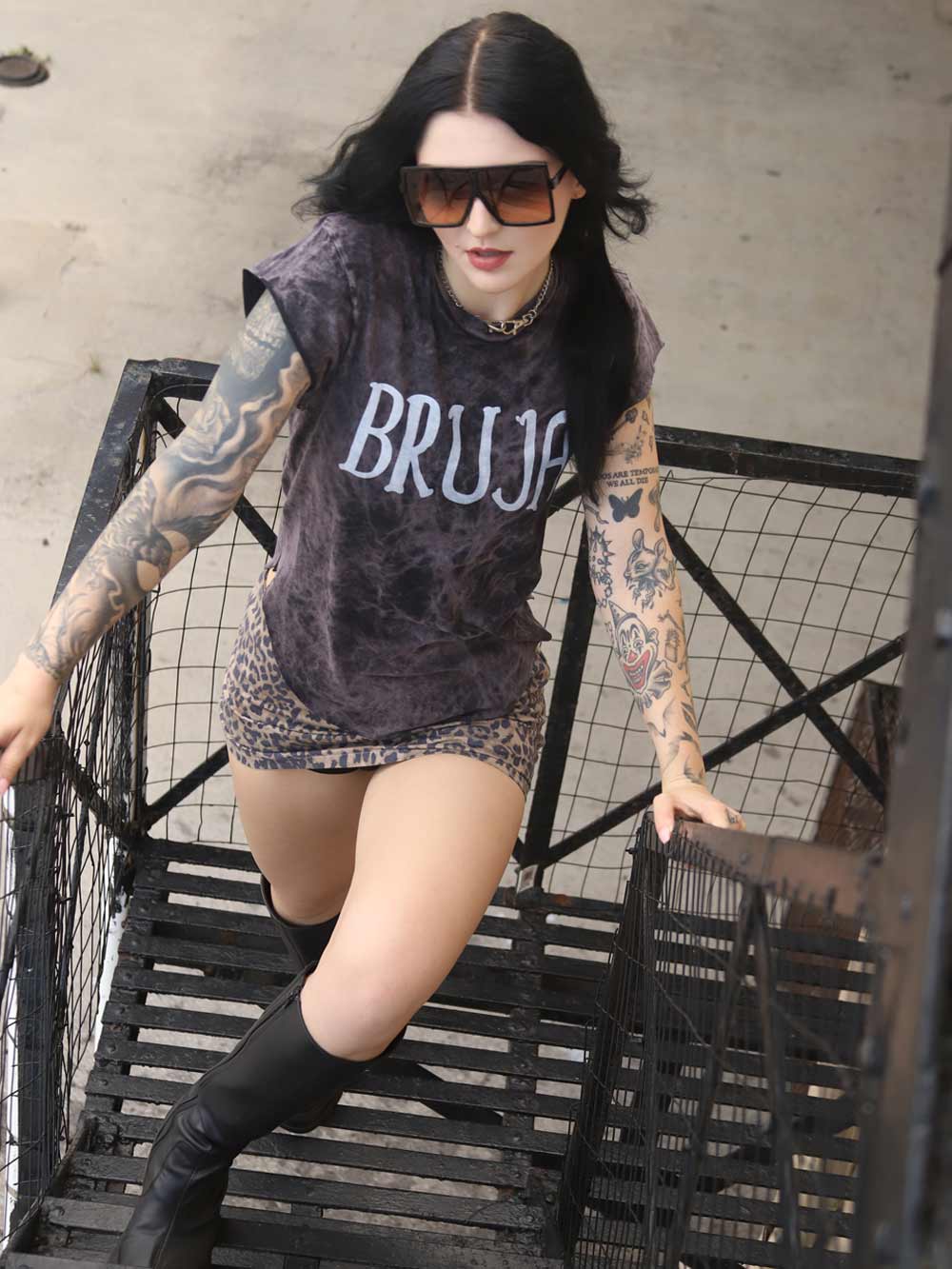 Person wearing a 'BRUJA' t-shirt with tattoos leaning against a metal staircase.