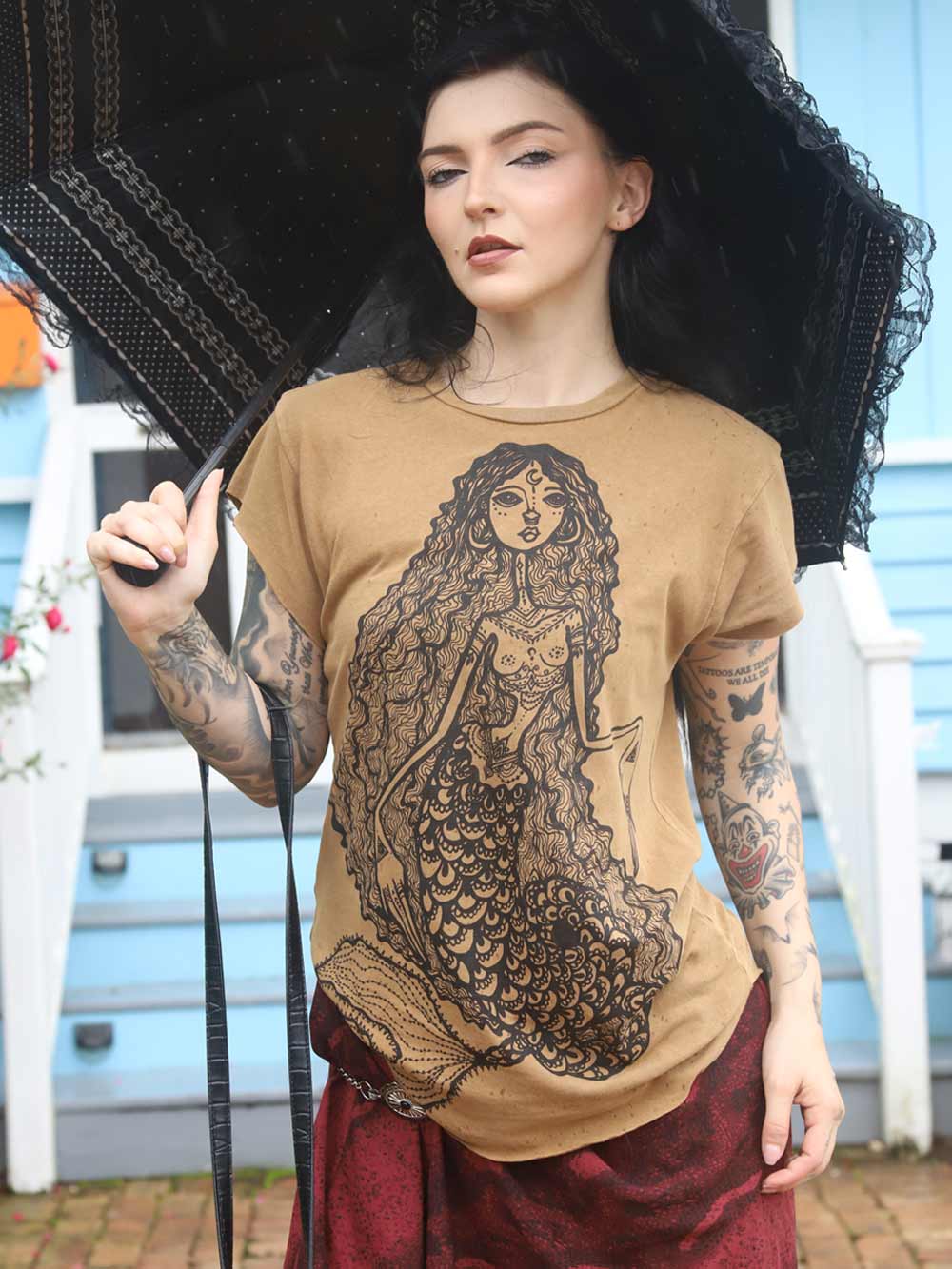 Woman wearing a tan t-shirt with a Mermaid graphic design, holding an umbrella.