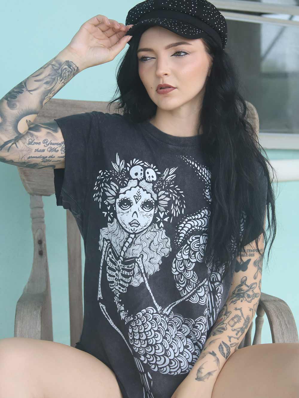 Woman wearing a black t-shirt with a white graphic design of a sugar skull mermaid, sitting on a wooden chair.