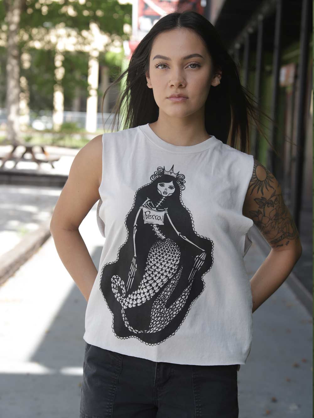 A woman wearing a white crop with a black graphic of a mermaid skeleton wearing a shirt with the word Perra on it.
