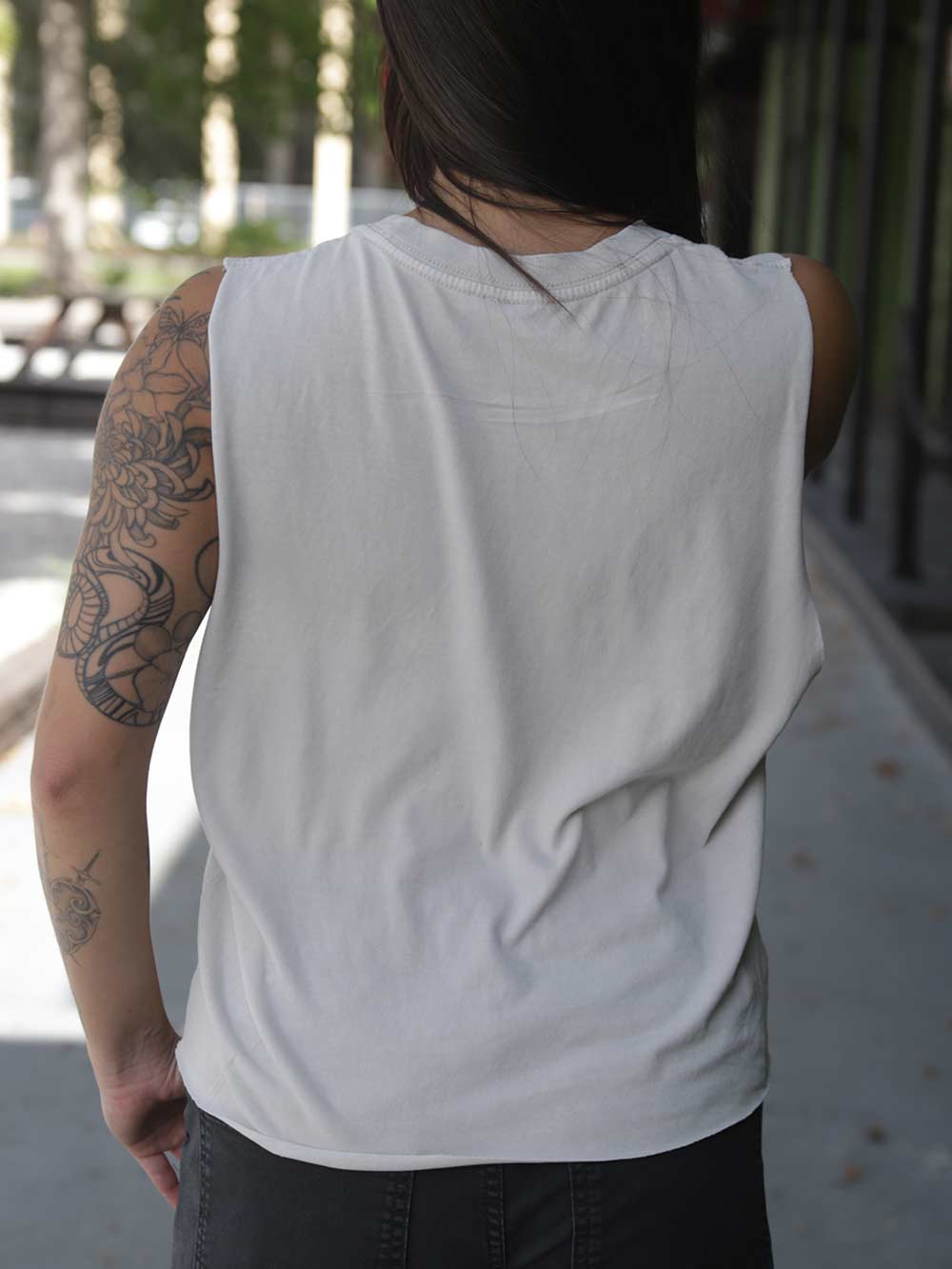 A person viewed from behind wearing a sleeveless white tank top and dark pants, standing outdoors on a paved walkway with trees and a building in the background.