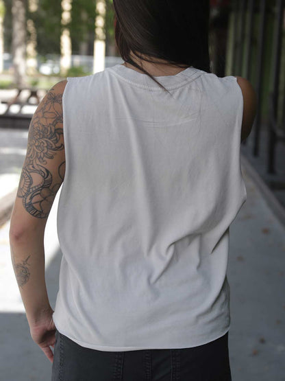 A person viewed from behind wearing a sleeveless white tank top and dark pants, standing outdoors on a paved walkway with trees and a building in the background.