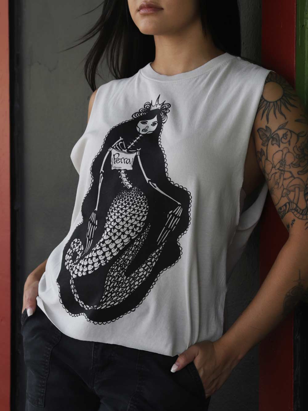 A woman wearing a white crop with a black graphic of a mermaid skeleton wearing a shirt with the word Perra on it.