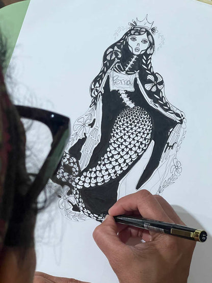 A hand holding a black pen draws a detailed black ink illustration of a mermaid with a crown on white paper. The artist's hand and part of their head with glasses are visible in the foreground.