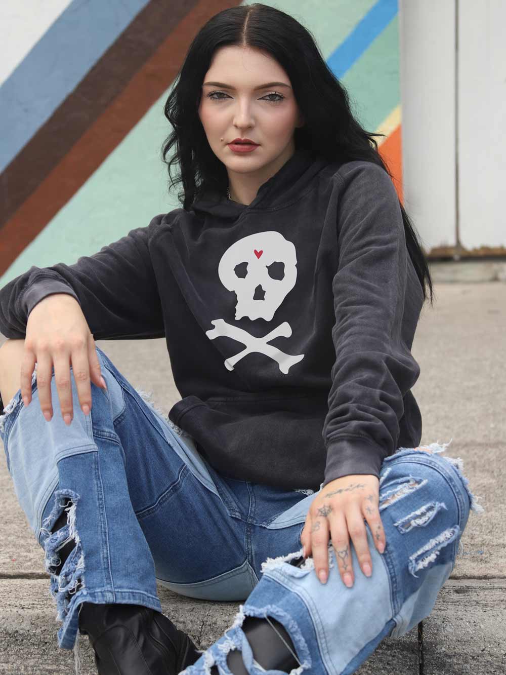 Woman wearing a black hoodie with a skull design and ripped jeans sitting on steps.