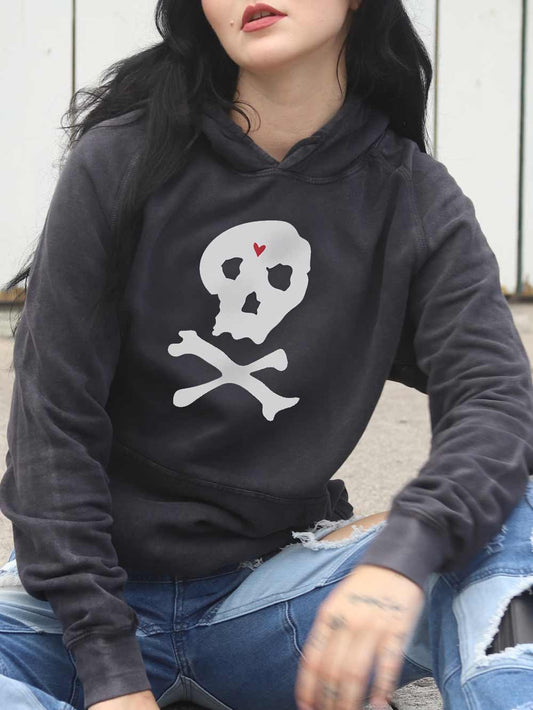 Woman wearing a black hoodie with a skull and crossbones design, sitting on a concrete surface.