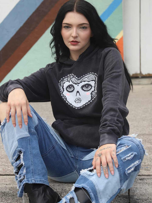 Model wearing a black hoodie with a white heart face graphic design and ripped jeans sitting on a concrete surface.