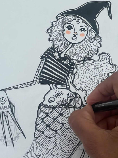 Hand-drawn illustration of a mermaid-like character with a witch's hat and tattooed arm, being completed by a hand holding a pen.