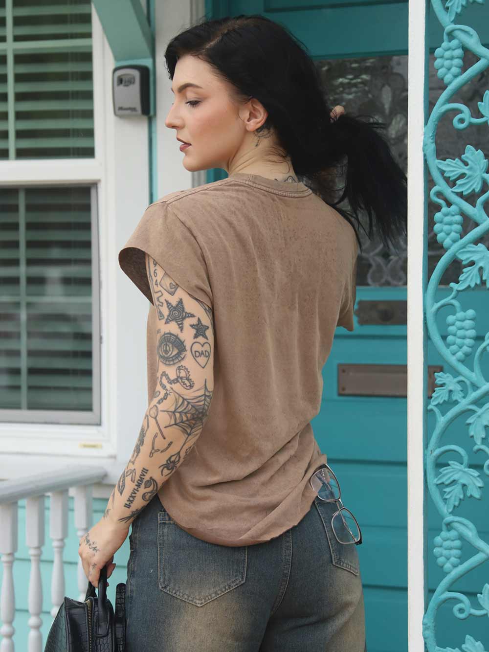 Woman with tattoos on her arm wearing a brown t-shirt and jeans, standing in front of a teal door.