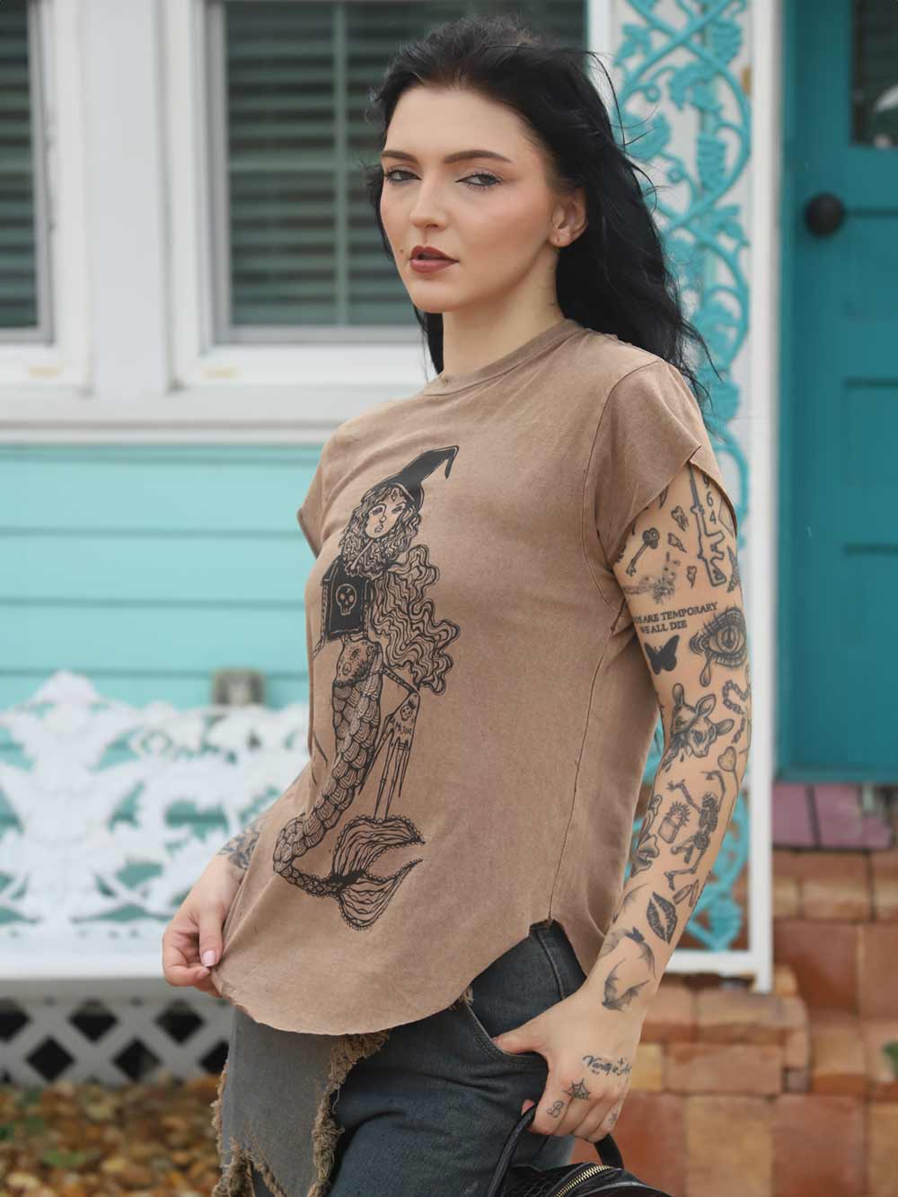 Woman wearing a brown t-shirt with a mermaid graphic design, standing in front of a teal house.