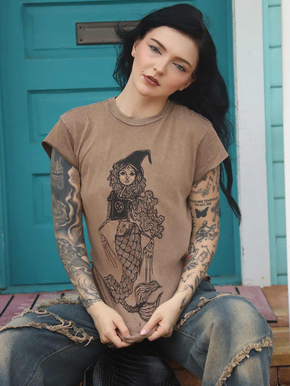 Woman wearing a brown t-shirt with a Mermaid graphic design, sitting in front of a teal door.