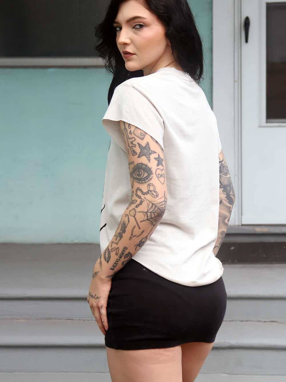 Woman with tattoos wearing a white sleeveless top and black shorts standing outdoors.
