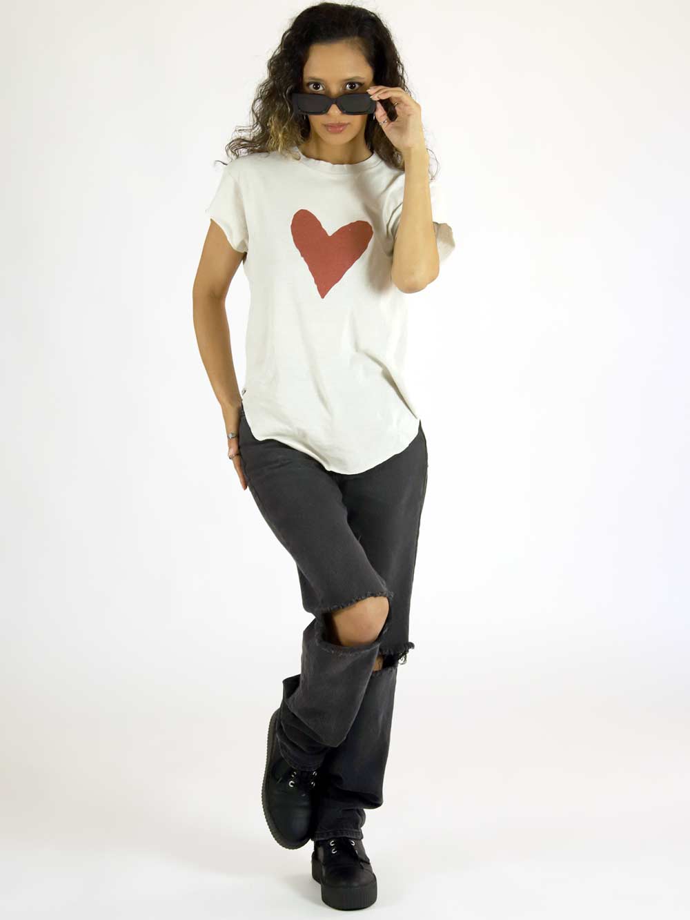 Woman wearing a white t-shirt with a red heart design, black pants, and black shoes on a white background, holding sunglasses.