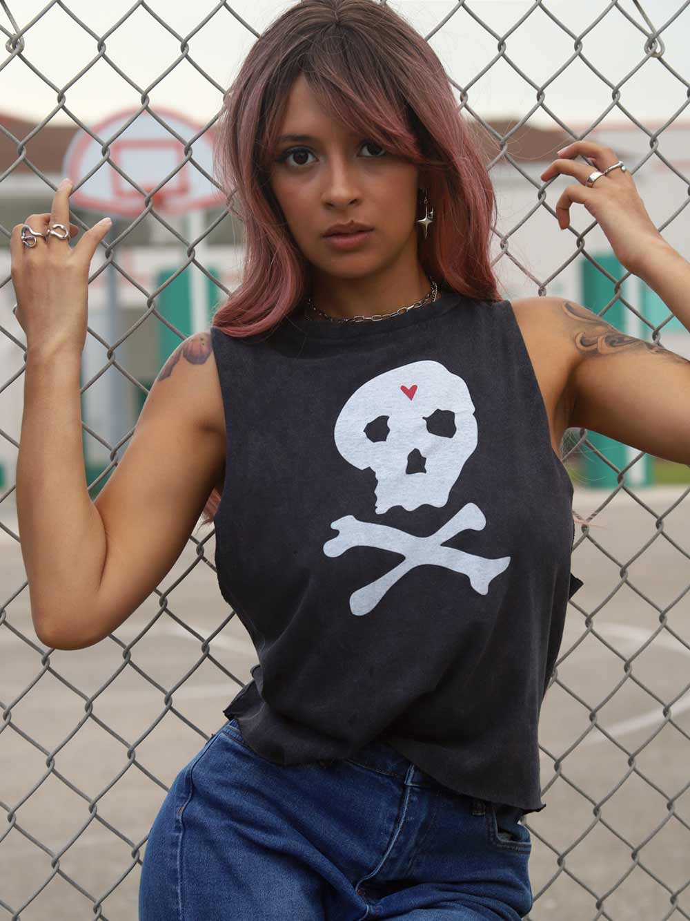 Lady wearing a black sleeveless shirt with a skull and crossbones design, standing in front of a chain-link fence.