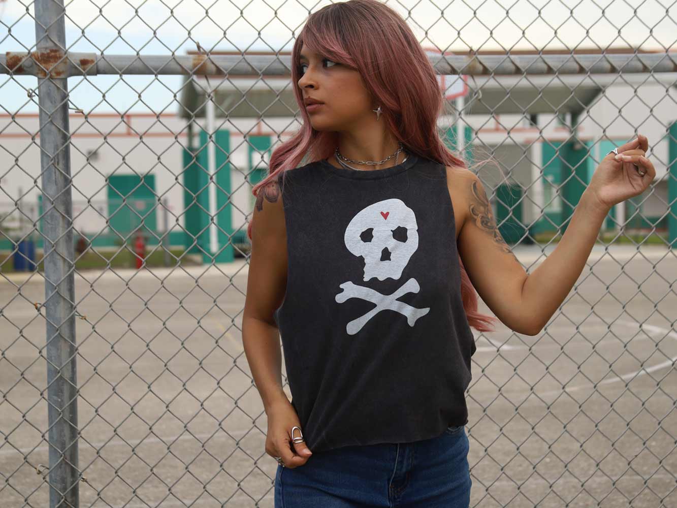 Person wearing a black tank top with a skull and crossbones design in front of a chain-link fence.