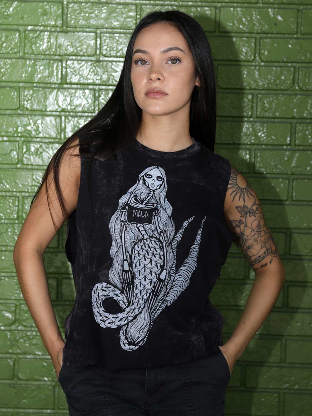 A woman with long dark hair and tattoos stands in front of a green brick wall, wearing a black sleeveless muscle tank top with a large mermaid graphic on the front.