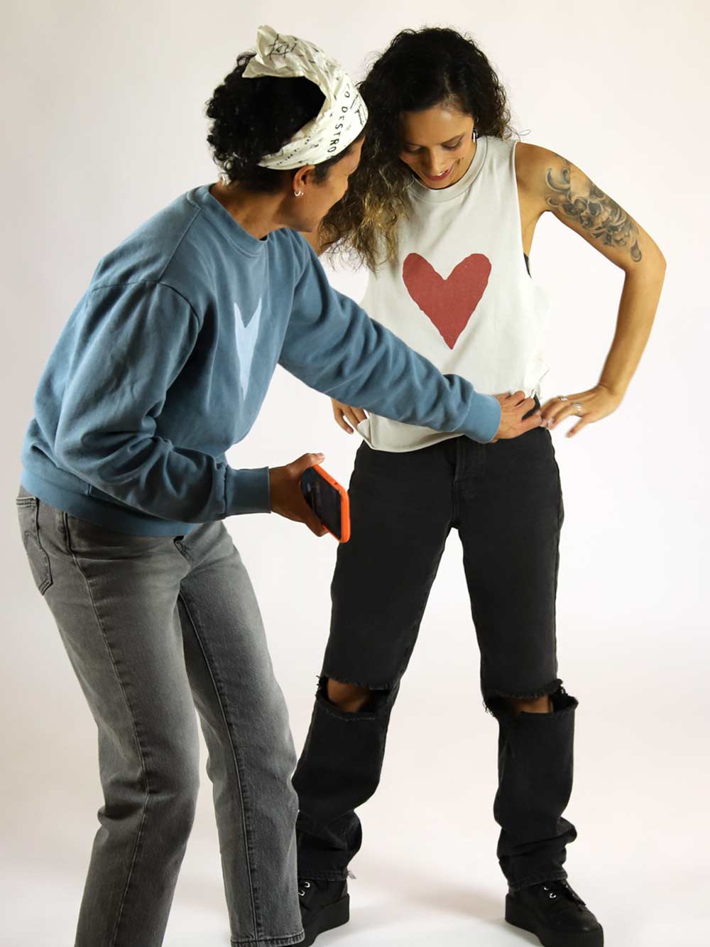 Photoshoot BTS, one wearing a blue sweater and gray pants, the model wearing a white tank top with a red heart and black pants.