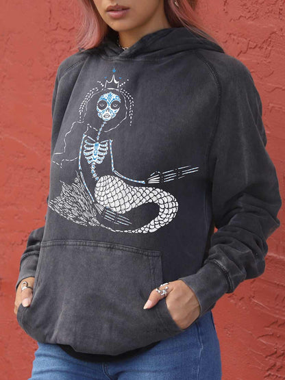 lady wearing a black hoodie with a skeleton mermaid design against a red wall.