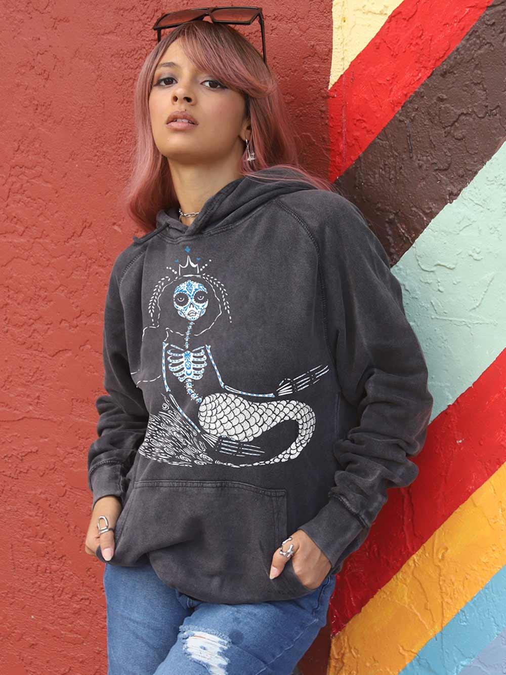 Woman wearing a black hoodie with a mermaid graphic design against a colorful wall.