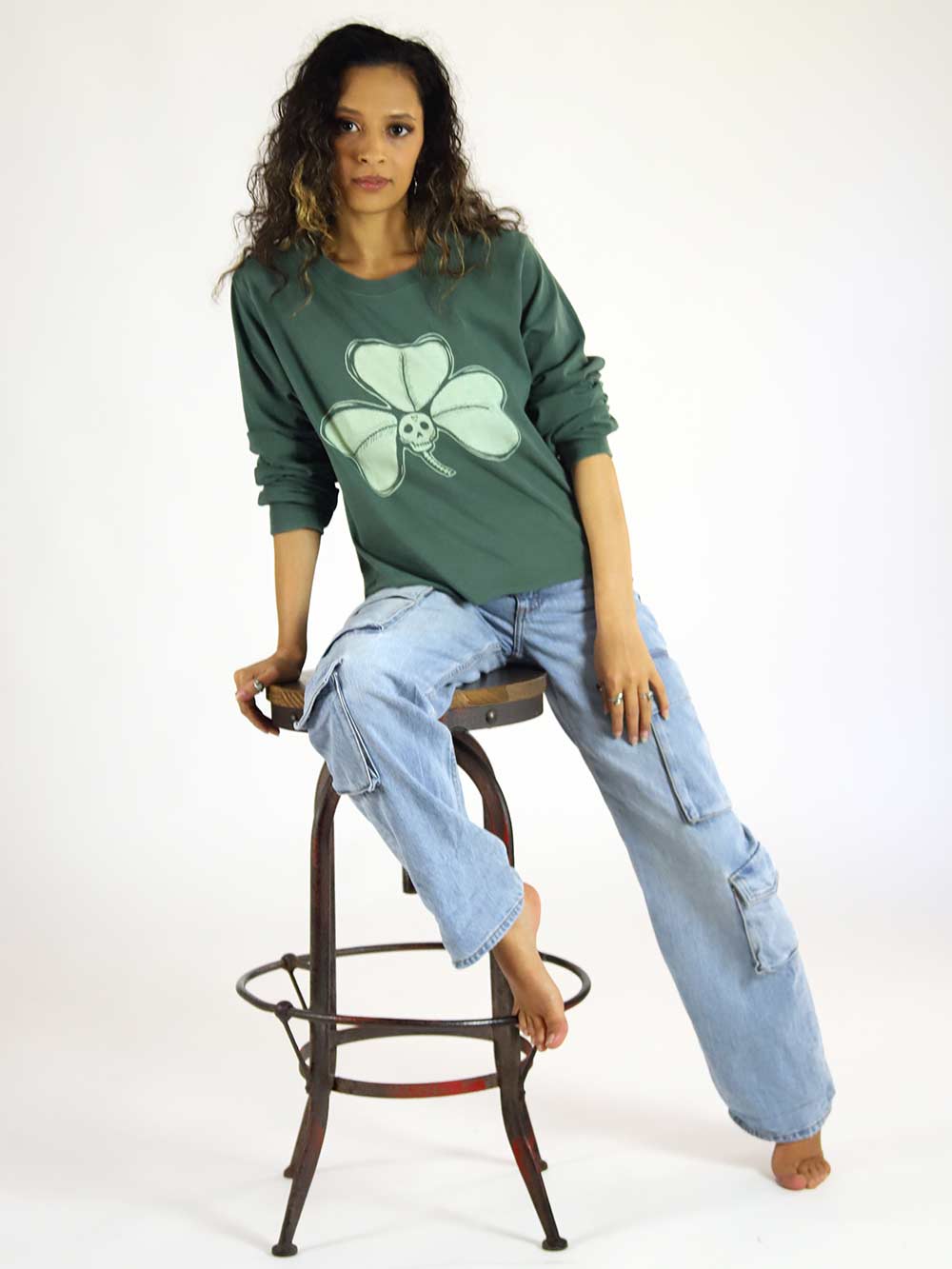 Woman wearing a green long sleeve shirt with a clover design, sitting on a stool against a white background
