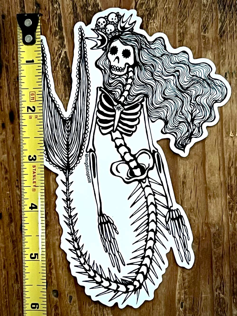 Skeleton Mermaid sticker with long hair and a broomstick on a wooden surface with a ruler for scale.