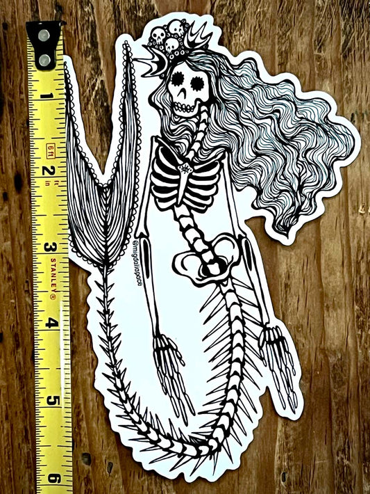 Skeleton Mermaid sticker with long hair and a broomstick on a wooden surface with a ruler for scale.