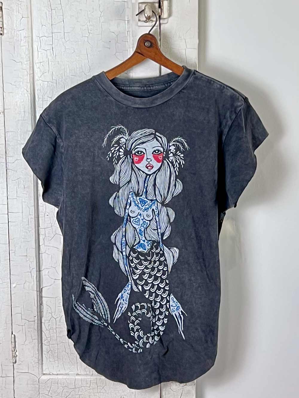 Dark gray t-shirt with a mermaid graphic on a wooden hanger against a white wall.