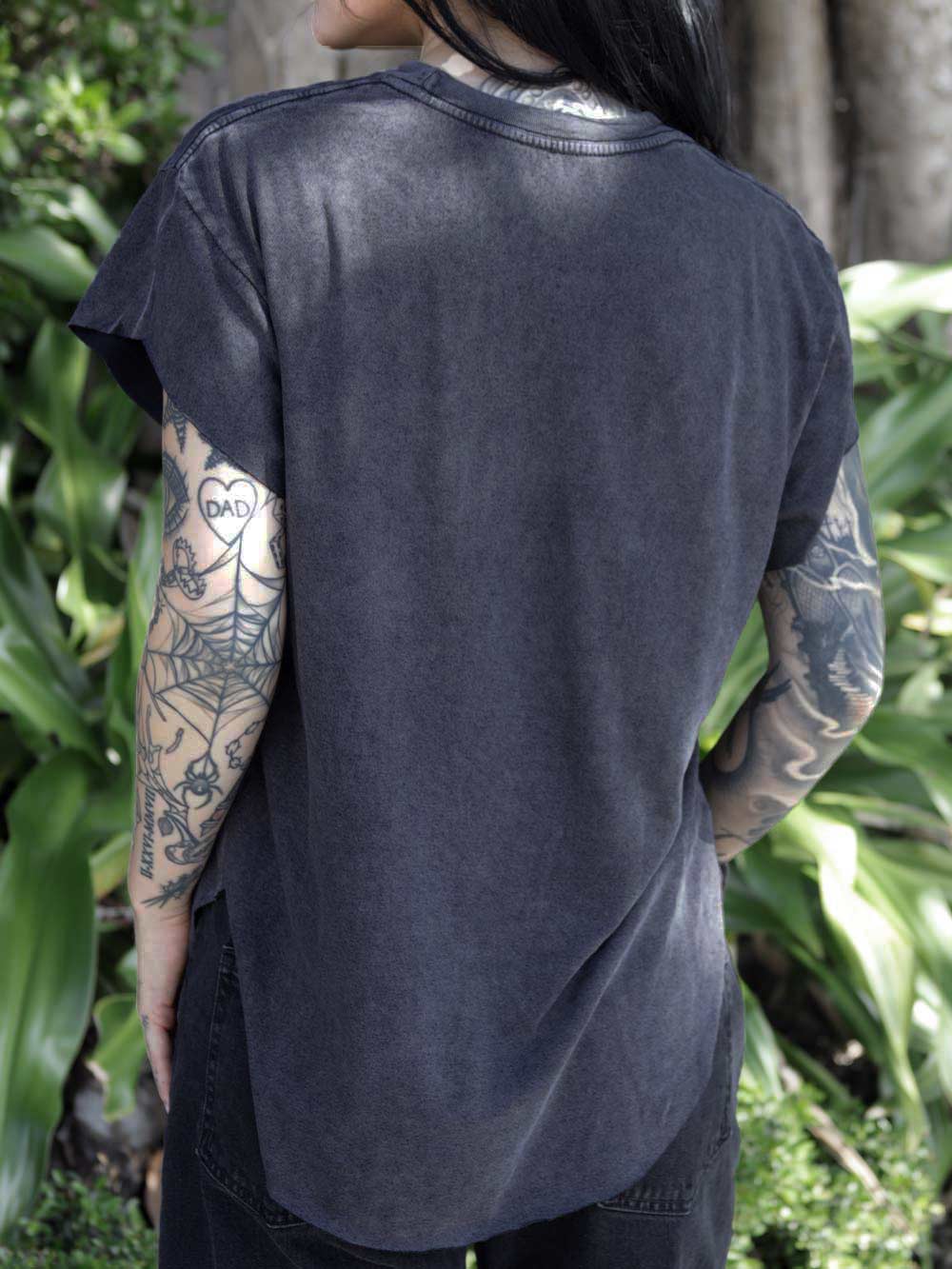 Person wearing a black t-shirt with tattoos on arms standing in front of green foliage