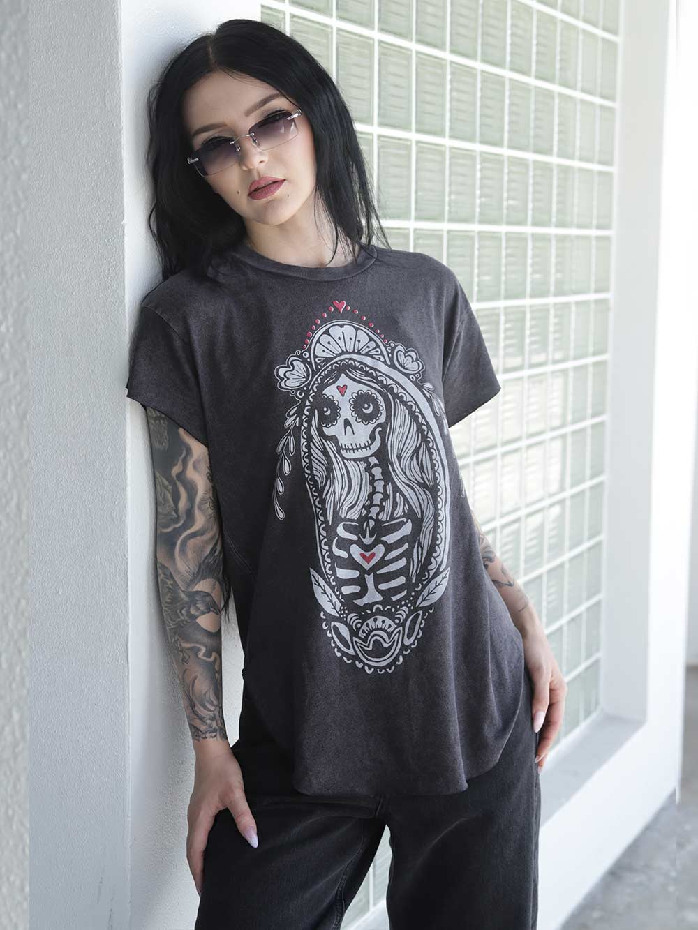 Woman wearing a black t-shirt with a sugar skull Madonna Graphic, standing against a glass wall.
