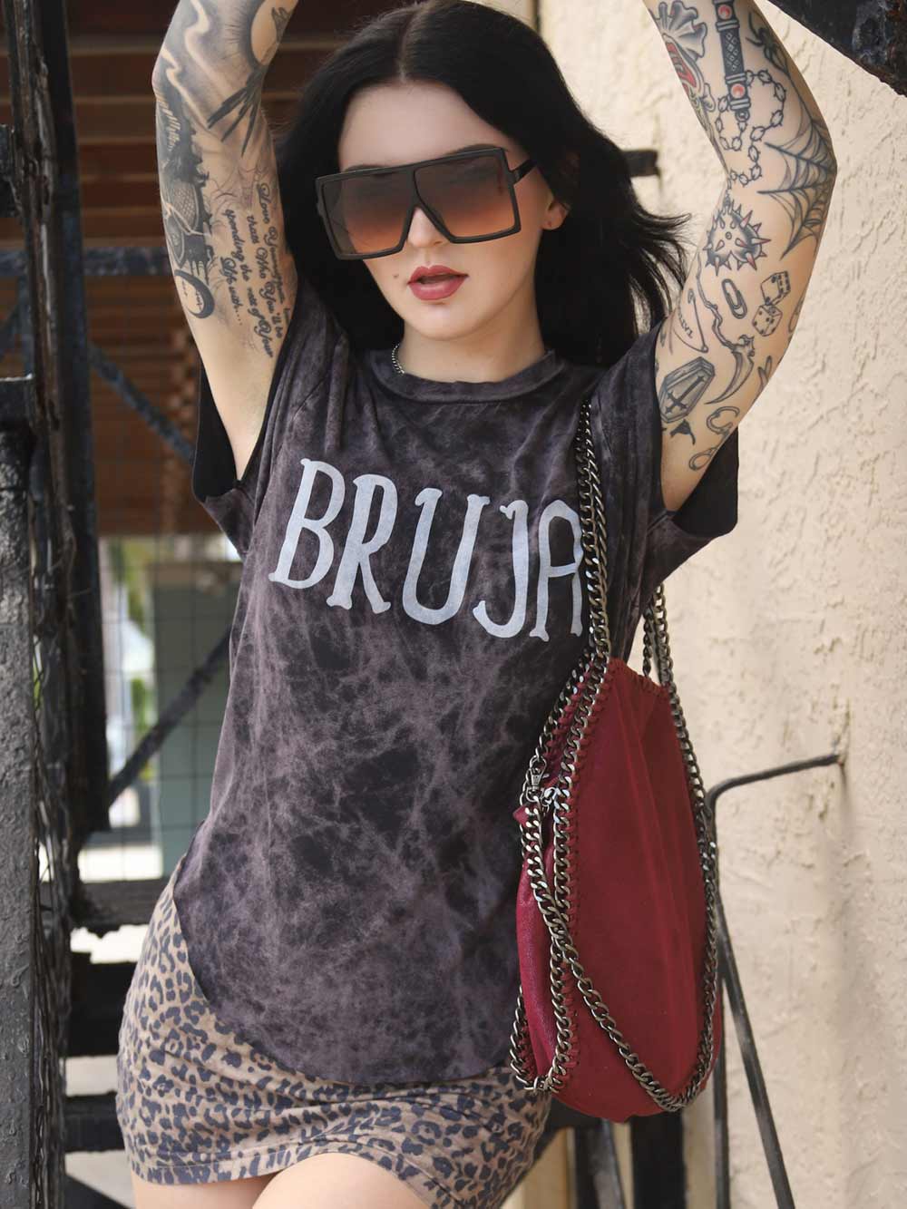 Person wearing a 'BRUJA' t-shirt with tattoos and a red bag.