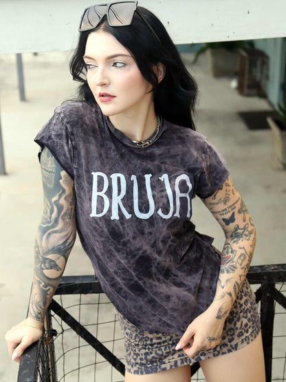 Person wearing a 'BRUJA' t-shirt with tattoos on arms, standing outdoors.