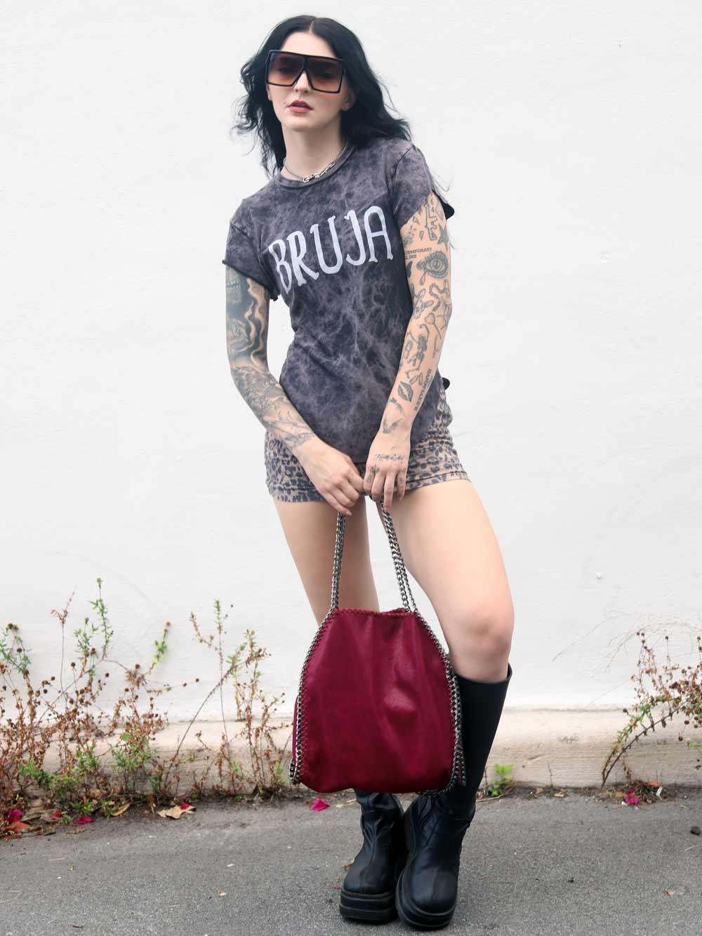 Woman wearing a 'BRUJA' shirt holding a red handbag against a plain background