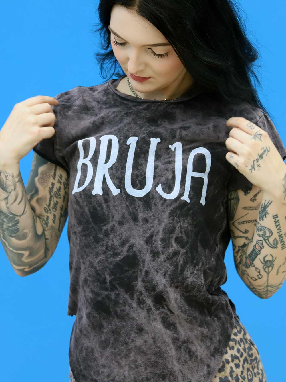 Person wearing a black tie-dye t-shirt with 'BRUJA' text against a blue background