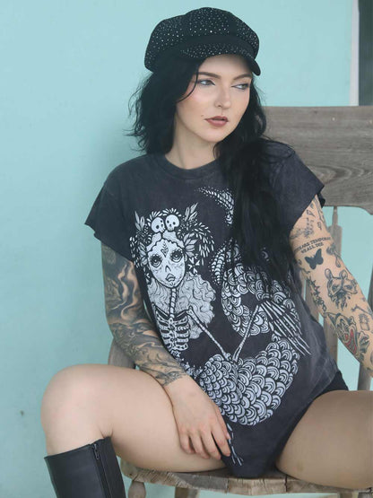 Woman wearing a black sleeveless shirt with a sugar skull mermaid graphic design, sitting in rocking chair.