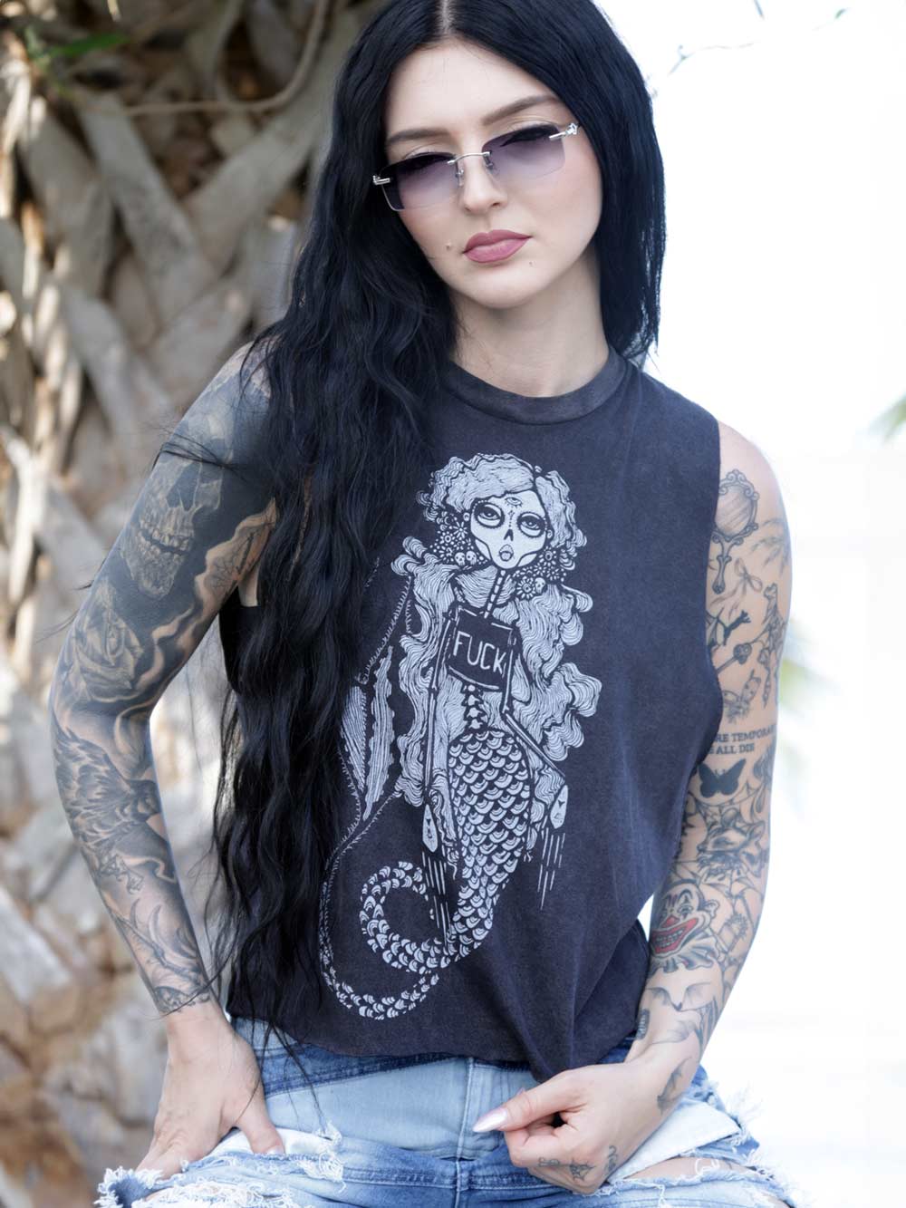 Woman wearing a sleeveless black top with a graphic design of a skeleton Mermaid wearing a shirt with the word fuck on it, sitting outdoors.
