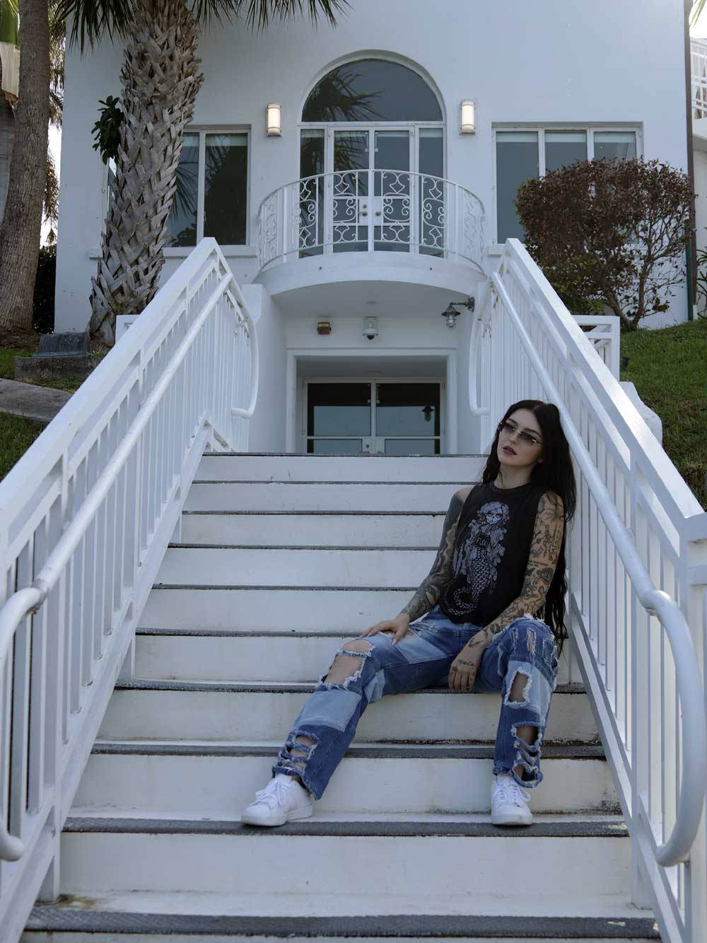 Person sitting on white steps in front of a white house with palm trees. Migdalia