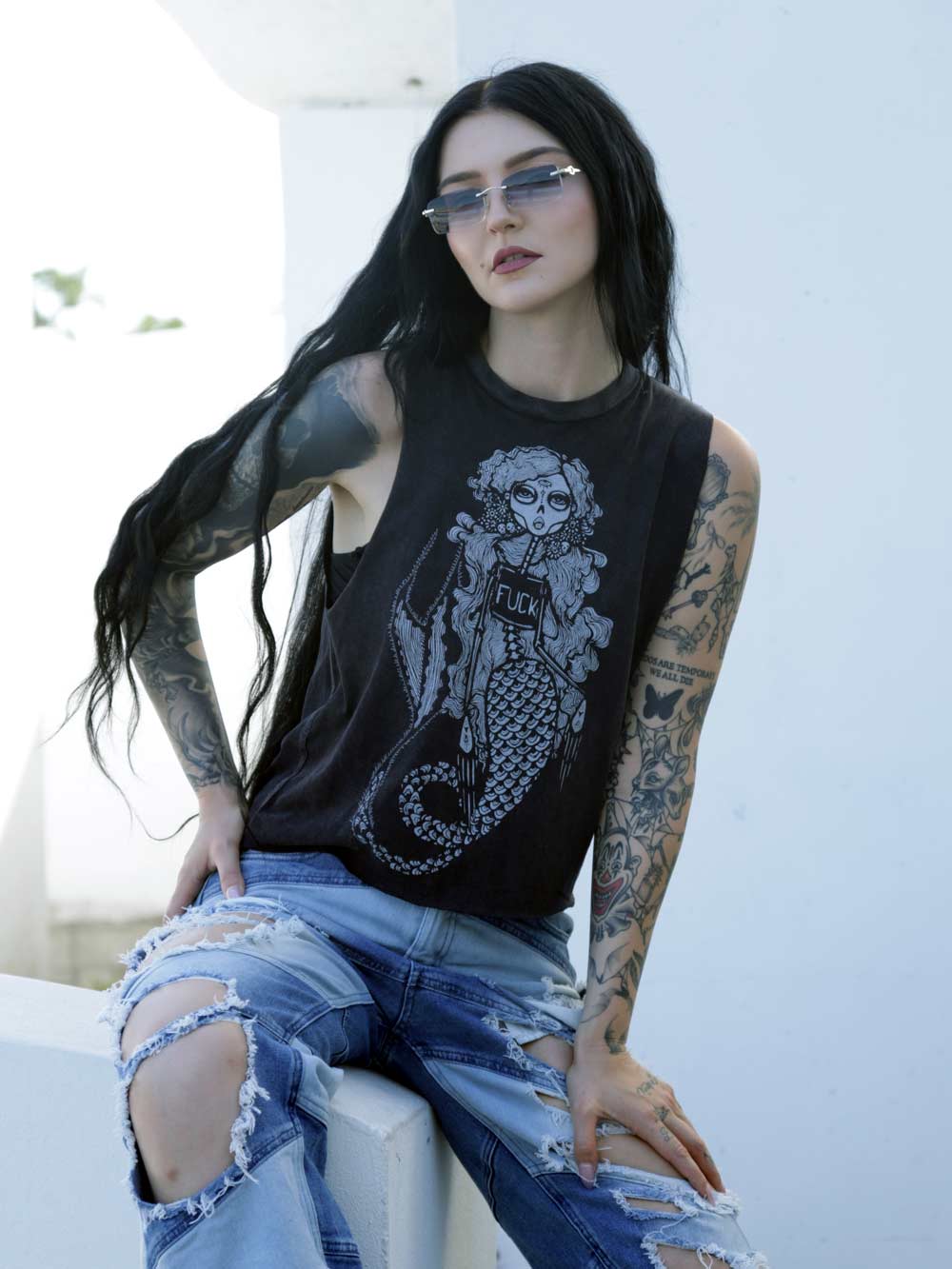 Woman wearing a sleeveless black top with a graphic design of a skeleton Mermaid wearing a shirt with the word fuck on it, sitting outdoors.